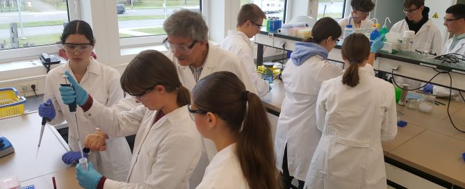 Auf diesem Bild sind Schülerinnen und Schüler in weißen Kitteln zu sehen, die im Labor arbeiten.